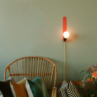 Lámpara de pared Skipper en color coral transparente delante de una pared verde oliva, junto a un sillón de ratán.